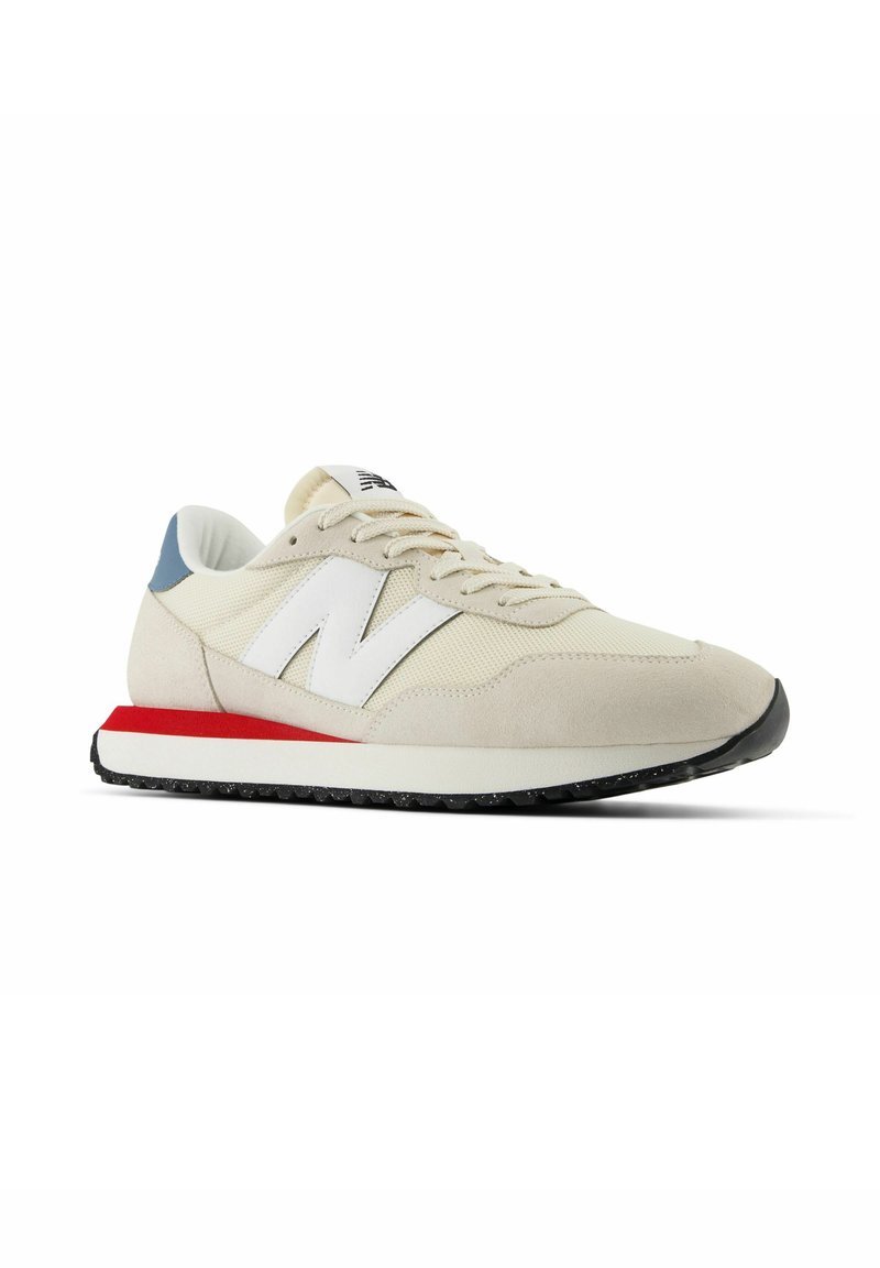 Beige Sport-schuh aus Netzmaterial und Wildleder, mit einem weißen "N"-Logo, blauen Akzenten an der Ferse, rotem Mittelsohlenstreifen und schwarzem Gummisohle.