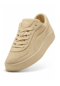 Zapatilla de ante beige con una suela de goma texturizada, diseño con cordones y un sutil logo de Puma en la lengüeta, que muestra curvas y costuras suaves.