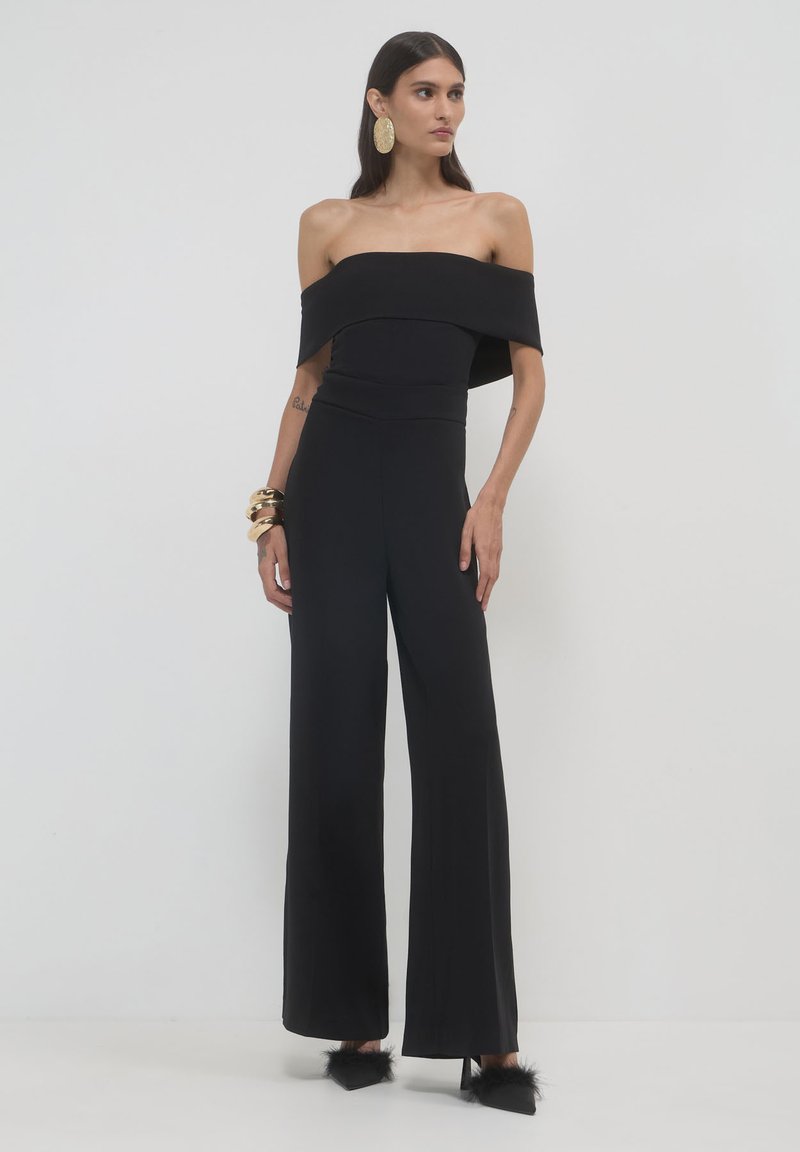 Jumpsuit nero scollato con spalle scoperte, gambe ampie, corpetto aderente e texture liscia. Orecchini rotondi d'oro di notevole fattura e tacchi neri pelosi.