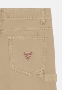 Beige bomullsbukser med et distinkt trekantet rødt og hvitt "GUESS" logo-patch på baklommen og rett snitt design.