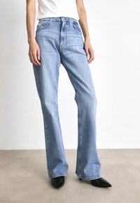 Vaqueros acampanados de denim azul claro con una cintura de altura media, un ligero desvanecimiento y detalles cosidos. Combinados con zapatos negros de punta afilada.