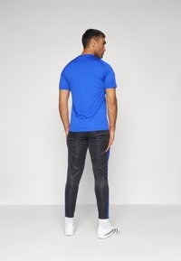 Blaues Sport-T-Shirt mit kurzen Ärmeln; kombiniert mit schwarzen, figurbetonten Sporthosen mit blauen und orangefarbenen Seitenschlitzen. Weiße Sportschuhe.