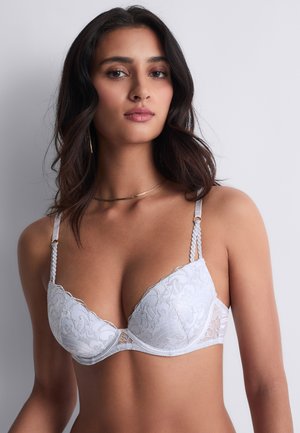 Femme aux cheveux ondulés foncés portant un soutien-gorge en dentelle blanche, regardant devant elle avec une expression neutre sur un fond clair uni.