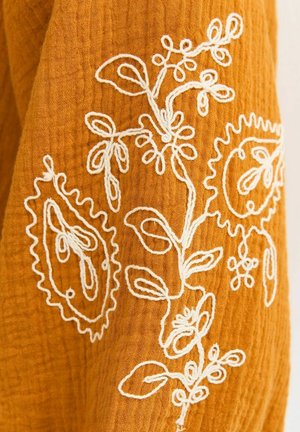 Mosterdkleurige stof met witte bloemenborduursels, bestaande uit bladeren, stelen en afgeronde paisley-achtige vormen in een gestructureerde weefstructuur.