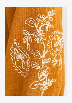 Tela color mostaza con bordado floral blanco que presenta hojas, tallos y formas redondeadas similares a cachemira en un tejido texturizado.
