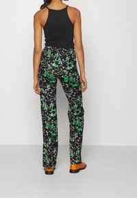 Top noir, mannequin portant un pantalon vert à motif floral sur fond noir, coupe slim, tissu lisse, associé à des chaussures marron.