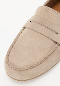 Beige mokasinikengät, joissa on pyöreä kärki, yksityiskohtaiset ompeleet ja koristeellinen nauha kengän päällä. Sileä pinta ja minimalistinen muotoilu.