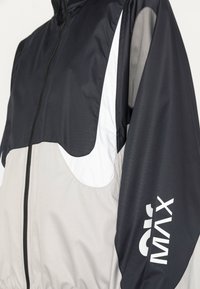 Vīriešu jaka ar melnbaltu krāsu shēmu, neilona materiālu, ar rāvējslēdzēju priekšpusē, elastīgu apakšmalu un baltu logotipa akcentu.