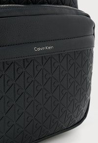 Juodas tekstūruotas kuprinės maišas su geometriniu raštu, lygiomis detalėmis ir metaliniu Calvin Klein logotipu priekyje.