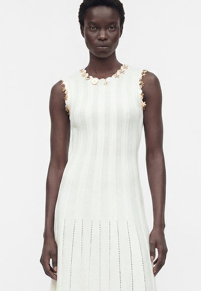 Femme portant une robe en maille blanche sans manches avec des motifs verticaux et de petites décorations florales autour de l'encolure et des emmanchures.