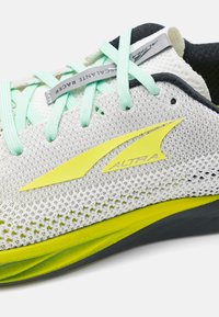 Chaussure de course légère avec une tige en mesh blanc, des lacets vert menthe, des accents jaunes et une semelle grise. Présente un design épuré et une bonne respirabilité.