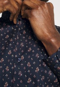 Svart skjorta med knappar som har ett upprepat blommönster i rött och grönt. Närbild som framhäver tygets struktur och knappdetaljer.