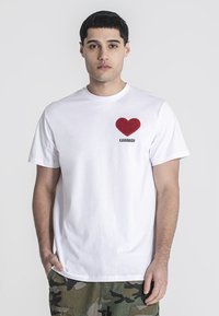 Camiseta de algodón blanca con un aplique de corazón rojo y el texto negro "KAVANAGH" en la parte izquierda del pecho. Diseño de mangas cortas y cuello redondo.