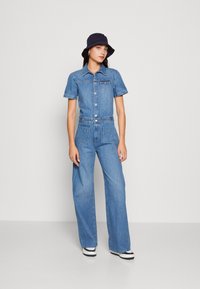 Jeans overall s kratkimi rokavi, ovratnikom, sprednjimi gumbi in dvema žepoma. Ohlapni hlačnice, svetlo modre barve, v kombinaciji z mornarsko modrim klobukom.