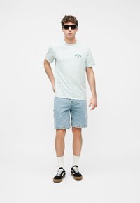 Νέος άνδρας που φοράει μαύρα γυαλιά ηλίου, ανοιχτό μπλε μπλουζάκι Rip Curl, τζιν σορτς, λευκές κάλτσες και μαύρα αθλητικά παπούτσια, στέκεται μπροστά σε λευκό φόντο.