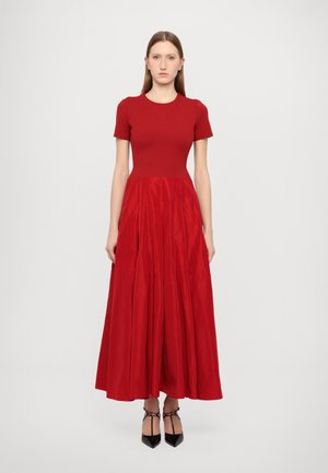Red dress with a fitted bodice and flared skirt. Short sleeves, made of smooth fabric, featuring a textured, pleated bottom. Black heels.
