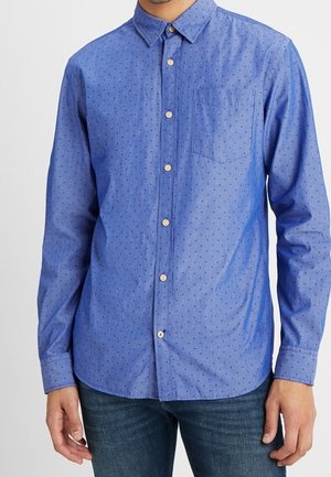 Heren overhemd met lange mouwen in blauw met kleine donkere stippen, borstzak en klassieke kraag, gedragen met een blauwe spijkerbroek.