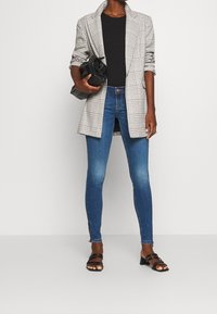 Persona vistiendo un blazer de cuadros, una blusa negra, jeans ajustados azules, tacones negros de tiras y sosteniendo un bolso de mano texturizado negro.
