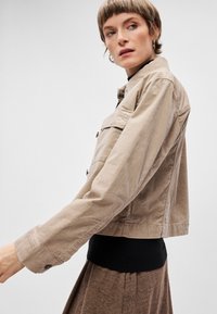 Veste courte de couleur beige clair avec un devant à boutons, deux poches poitrine et des manches longues, associée à une taille en tricot foncé.