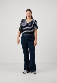 Stripet kortermet topp i marineblått og hvitt, i kombinasjon med mørkeblå vide jeans. Modellen holder en svart veske og har på seg sølvfargede joggesko.