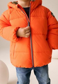 Veste matelassée orange vif avec capuche, dotée d'une fermeture éclair frontale et d'un design matelassé. Associée à un jean bleu, affichant un look décontracté.