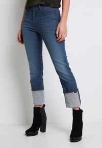 Jeans de mezclilla azul ajustados con puños en gris claro doblados, combinados con botines de ante negros con tacón. Textura suave, diseño de tiro medio.
