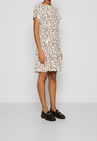 Vestido de manga corta en crema con un patrón en espiral en rojo, negro y blanco, de corte holgado y con dobladillo a la altura de la rodilla. Combinado con mocasines negros.