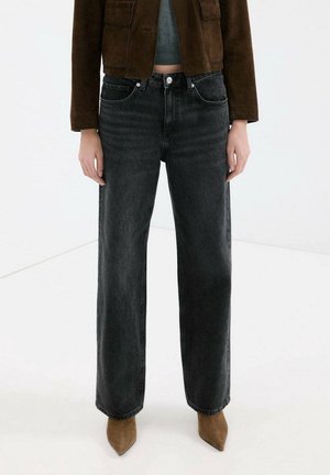 Persoon die zwarte jeans met hoge taille en wijde pijpen draagt, bruine suède spits toelopende laarzen, een korte bruine jas en een grijze top tegen een witte achtergrond.