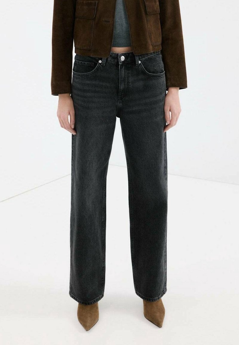 Personne portant un jean noir taille haute à jambes larges, des bottes pointues en daim marron, une veste marron courte et un haut gris sur fond blanc.
