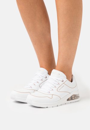 Personne portant des baskets blanche Skechers avec des accents beiges et des semelles à coussin d'air transparentes, se tenant sur une surface blanche.