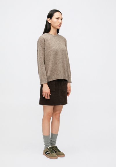 Femme debout de profil, vêtue d'un pull beige, d'une jupe marron foncé avec poches, de chaussettes grises et de baskets vertes, sur un fond blanc uni.