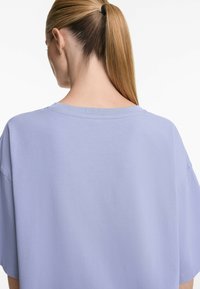 Světle fialové oversized tričko s kulatým výstřihem, jemnou texturou a krátkými rukávy, zobrazené z zadního pohledu a s hladkým, neformálním designem.