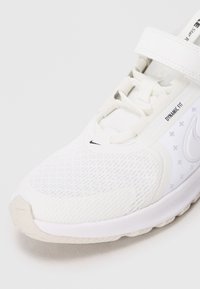 Chaussure de sport Nike blanche avec dessus en mesh, lacets et sangle Velcro, étiquette "Dynamic Fit" et semelle texturée.