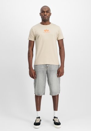Beige bomulds t-shirt med orange logo, parret med grå denim shorts. Modellen har sorte sneakers og hvide sokker på, stående foran en ensfarvet baggrund.