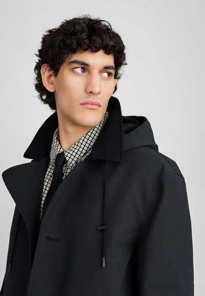 Young man with curly hair wearing a black coat over a patterned shirt and black tie, looking to the side against a plain background.