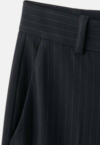 Primer plano de pantalones negros con rayas finas y verticales blancos, con cinturilla y trabilla para cinturón, mostrando tela plisada y rayas blancas verticales.