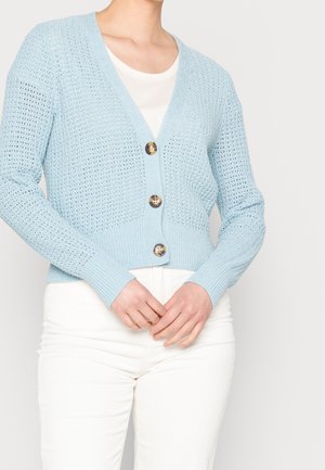Femme portant un cardigan en maille bleu clair avec trois boutons marron sur un haut blanc et un pantalon blanc, les mains jointes à la taille.