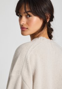 Lichtbeige trui met ribbelstructuur, ronde halslijn en verlaagde schouders. Heeft een ontspannen pasvorm en subtiele stikdetails.