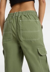 Grüne Cargo-Hose mit elastischem Bund, zwei Gesäßtaschen mit weißen Nähten und seitlichen Cargo-Taschen. Baumwollstoff-Textur.