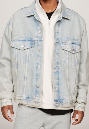 Mężczyzna w jasnoniebieskiej kurtce Tommy Jeans z denimu, noszący pod nią białą rozpinaną bluzę oraz jasne spodnie denimowe, pokazany od szyi do bioder.