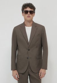 Mann in braunem Anzug mit weißem Hemd und dunkler Sonnenbrille, der vor einem schlichten hellen Hintergrund steht.