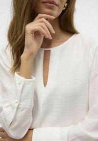 Blusa bianca a maniche lunghe con scollatura a chiave e polsini con bottoni. Il tessuto ha una finitura testurizzata, morbido e leggero.