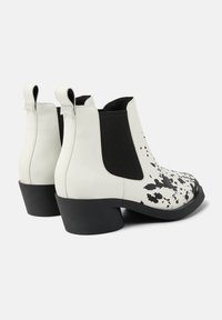 Botas de tobillo de cuero blanco con diseño de salpicaduras negras, paneles laterales elásticos y tacón grueso negro. Textura suave con lengüetas.