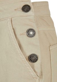 Prim-plan cu pantaloni din stofă bej, cu trei nasturi metalici inscripționați „Urban Classics” de-a lungul părții, aproape de cusătura buzunarului.