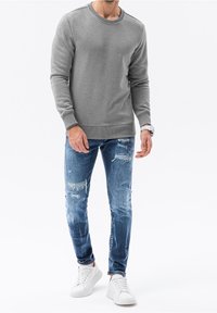 Uomo in piedi con indosso un felpa grigia a maniche lunghe, jeans blu strappati, sneakers bianche e un orologio bianco al polso sinistro.