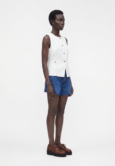 Femme portant un haut blanc sans manches avec des boutons, un short en jean bleu et des chaussures compensées marron, debout devant un fond clair uni.
