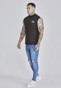 Black sleeveless top with a crew neckline, blue ripped skinny jeans, and white sneakers. The individual has visible tattoos on arms.