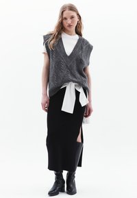Gilet grigio a maglia con scollo a V sopra una maglietta bianca, abbinato a una gonna nera con una spaccatura laterale e stivali neri al ginocchio.