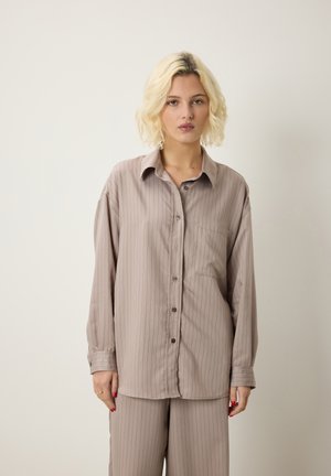Blonde vrouw met golvend haar, gekleed in een losse, beige gestreepte blouse en bijpassende broek, staand tegen een effen lichte achtergrond.