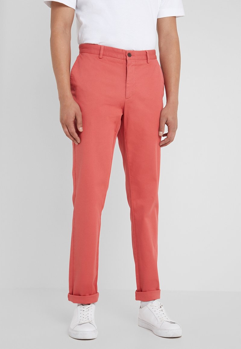 Personne portant un pantalon slim couleur corail avec des ourlets remontés, associé à des baskets blanches et une chemise blanche à manches courtes, sur un fond neutre.